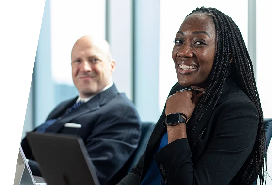 Two professionals in business attire sit at a table with laptops; the woman in the foreground is smiling while the man in the background looks on.