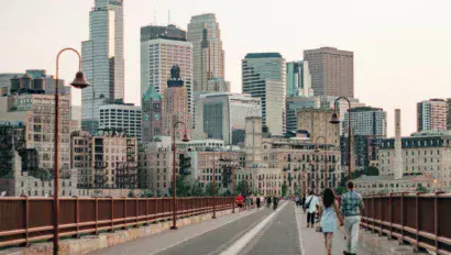 La gente camina por un puente peatonal con el horizonte moderno e histórico de una ciudad de fondo bajo un cielo despejado.
