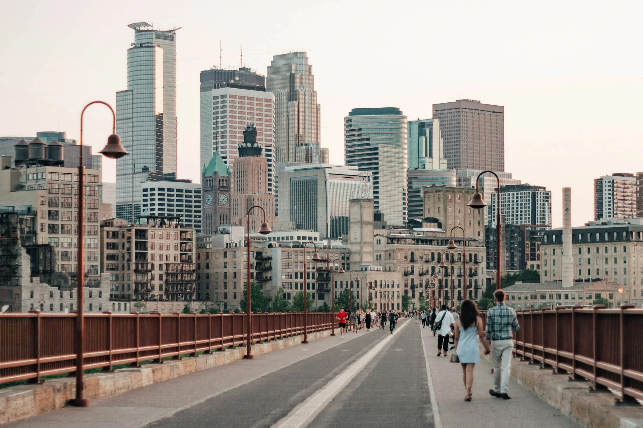 People walk along a pedestrian bridge with the modern and historic skyline of a city in the background under a clear sky.