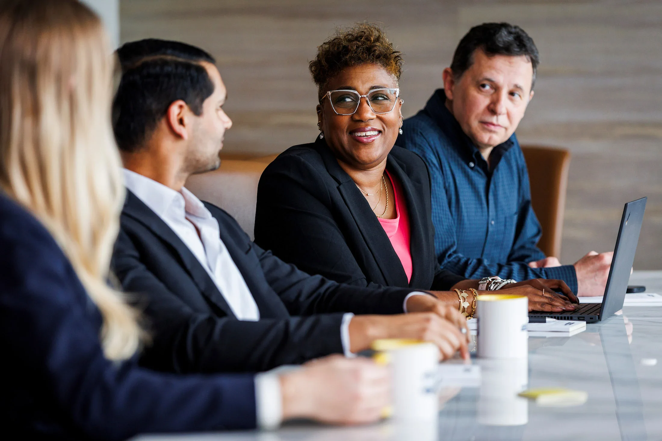 Four people sit at a conference table in a meeting, with one woman in glasses smiling and looking at the camera while others engage in conversation.