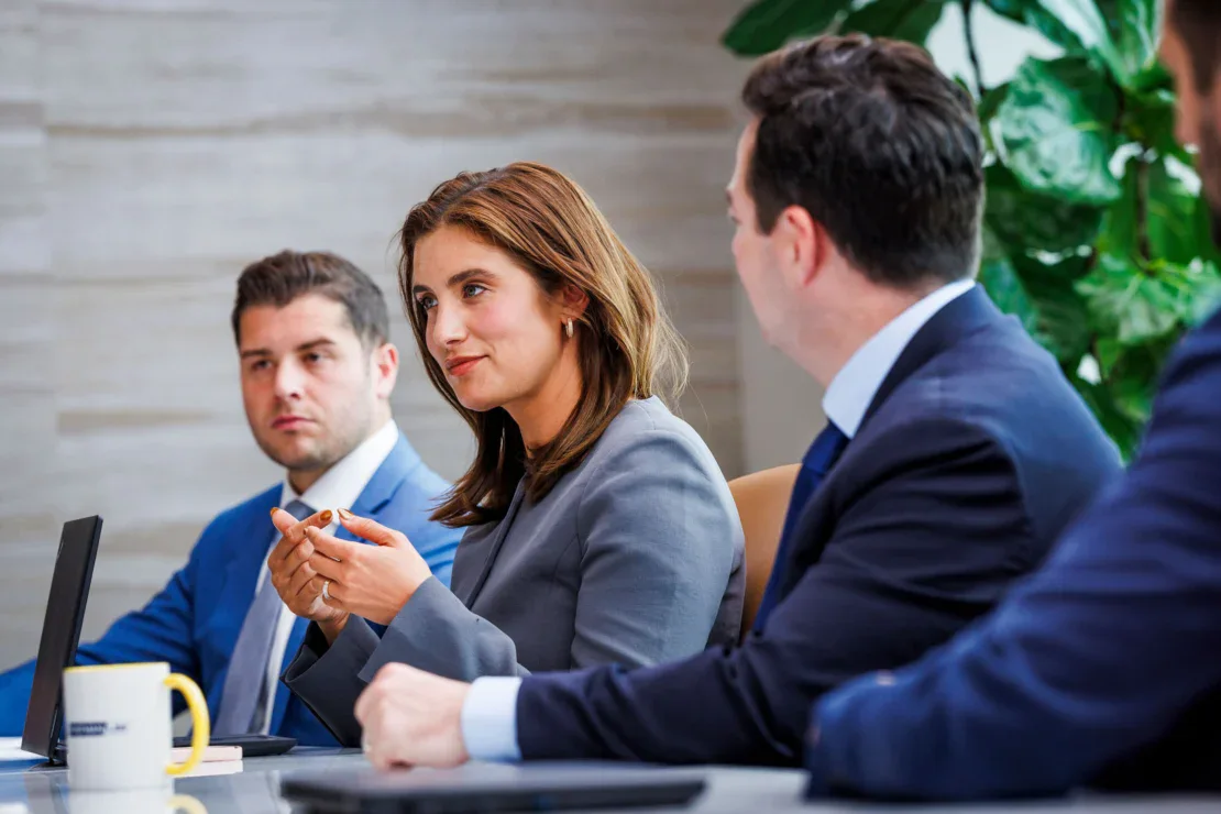 Tres personas en traje de negocios se sientan en una mesa de conferencias con ordenadores portátiles, participando en una reunión o discusión.