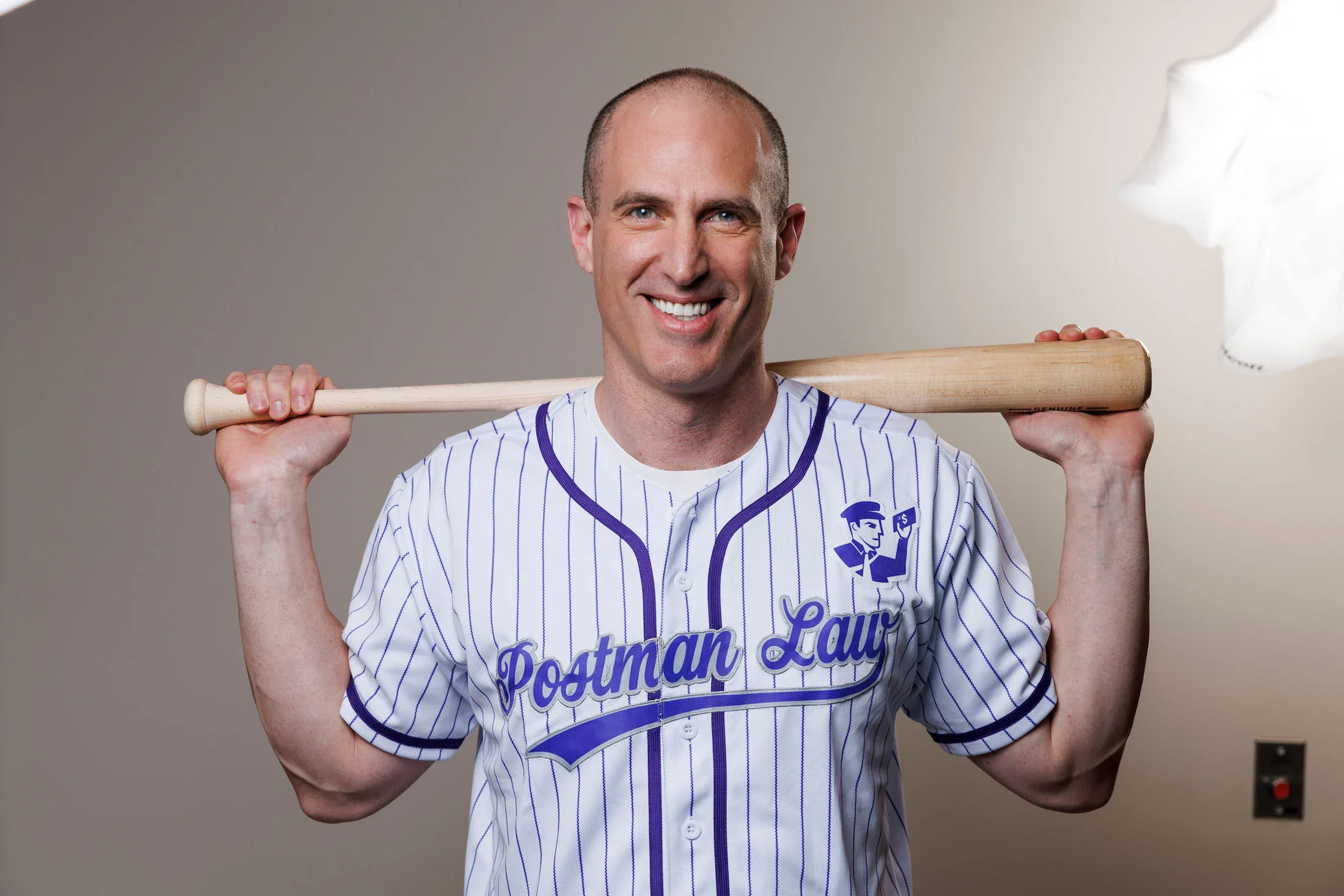 A man in a striped "Postman Law" baseball jersey smiles while holding a bat across his shoulders in front of a plain background.