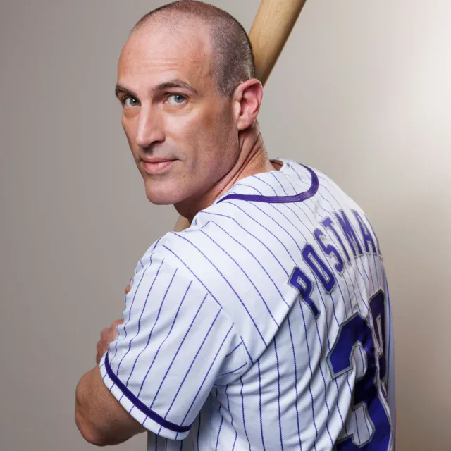 A man in a pinstriped baseball jersey with "POSTMAN" and the number 38 on the back holds a baseball bat over his shoulder and looks at the camera.