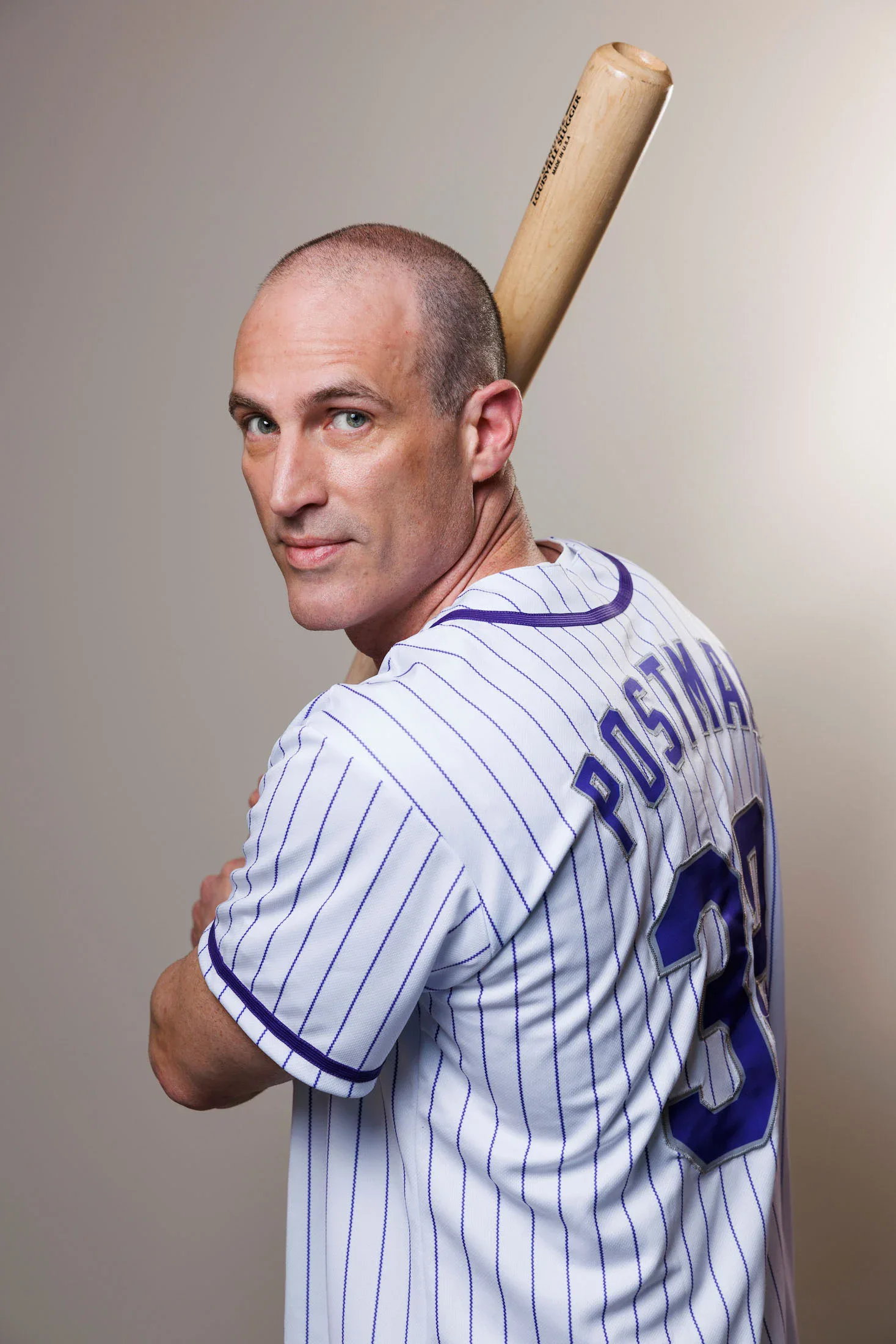 A man in a pinstriped baseball jersey with "POSTMAN" and the number 38 on the back holds a baseball bat over his shoulder and looks at the camera.