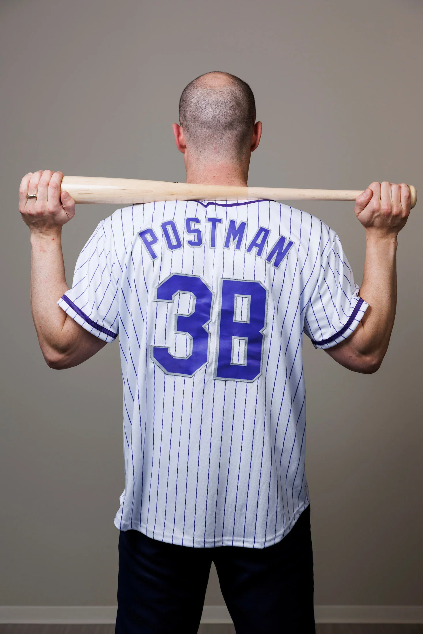 A person stands with their back to the camera, holding a baseball bat behind their shoulders. They wear a white pinstriped jersey with "POSTMAN" and "3B" on the back.