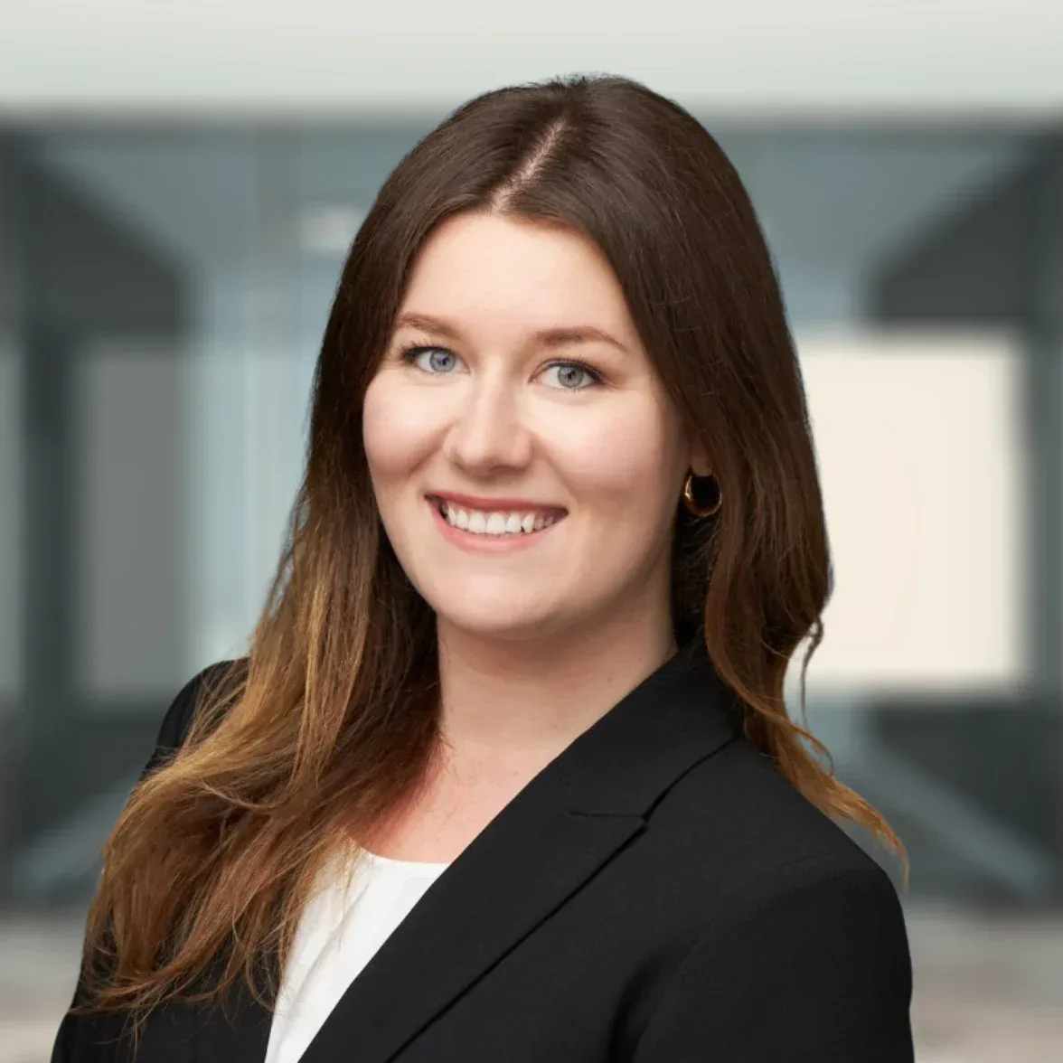 A woman with long brown hair wearing a black blazer and white top smiles in a modern office setting.