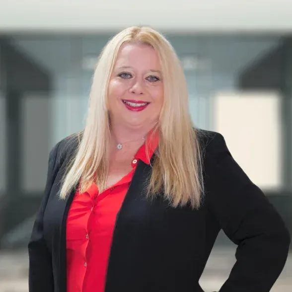 A woman with long blonde hair wearing a black blazer over a red blouse stands indoors, smiling at the camera.