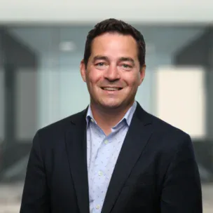 A man in a navy suit and light blue shirt is smiling, standing in front of a blurred office background.