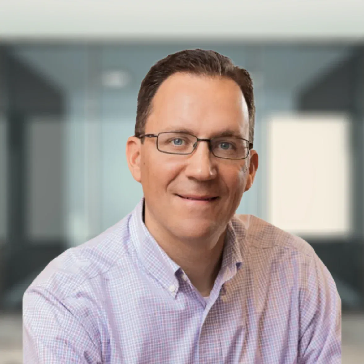 A man wearing glasses and a light purple checkered shirt smiles while seated in an office setting with glass walls.