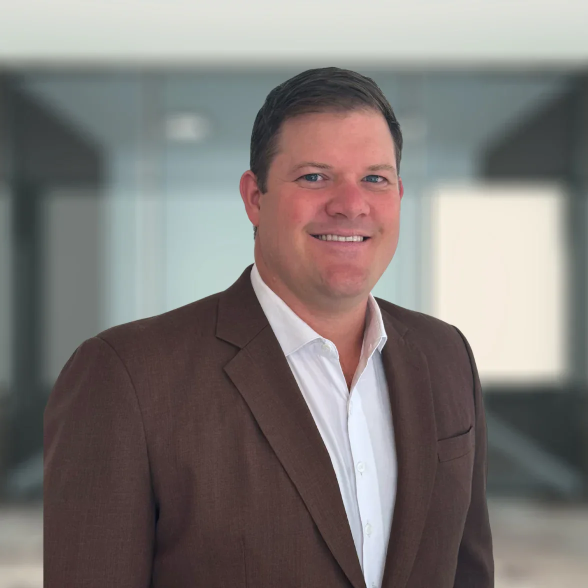 A man in a brown suit jacket and white shirt stands smiling in front of a blurred office background.