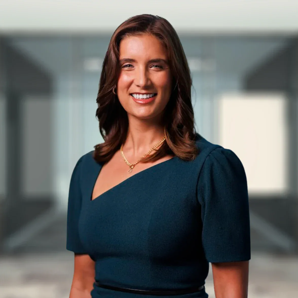 A woman with shoulder-length brown hair, wearing a dark teal dress and a gold necklace, stands in a modern office setting and smiles at the camera.