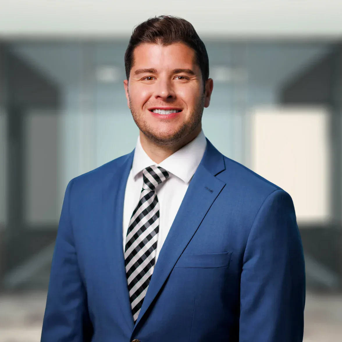 A man in a blue suit and checkered tie stands indoors, smiling at the camera with a blurred office background.