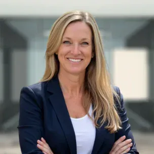 A woman with long blonde hair wearing a navy blazer and white top stands with arms crossed, smiling, in front of a modern glass building.