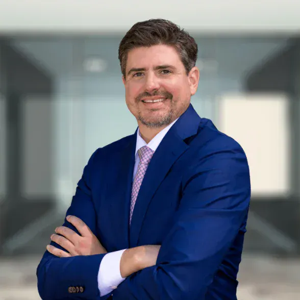 Man in a blue suit and light pink tie stands with arms crossed, smiling, in front of a blurred modern office background.