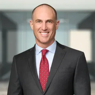 A man in a dark suit, light blue shirt, and red patterned tie smiles at the camera against a blurred office background.