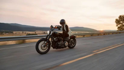 Person wearing a helmet rides a black motorcycle on a paved highway at sunset, with hills and open fields in the background.