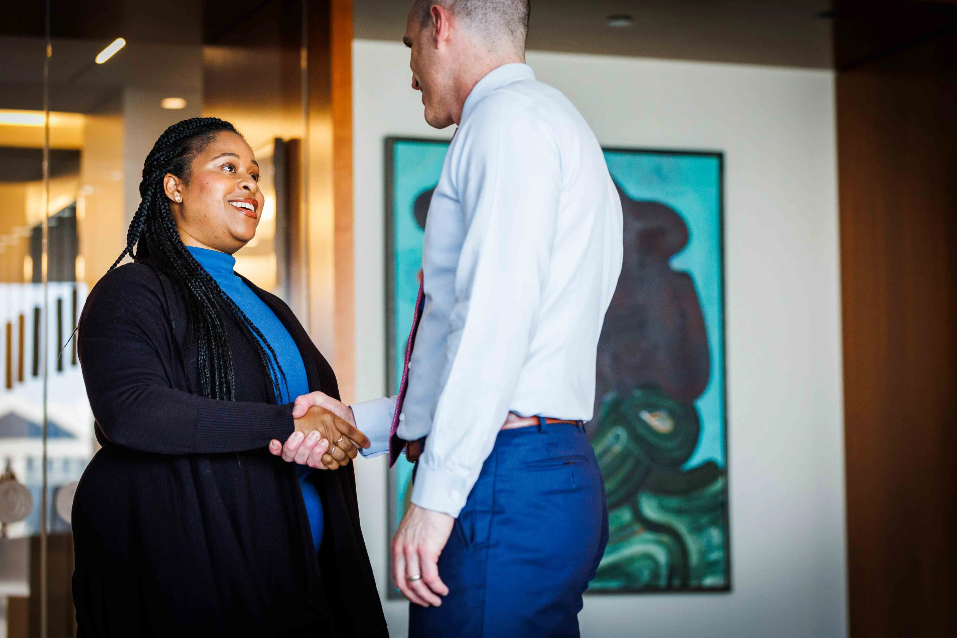 Two people in business attire shake hands and smile in a modern office setting with artwork on the wall in the background.