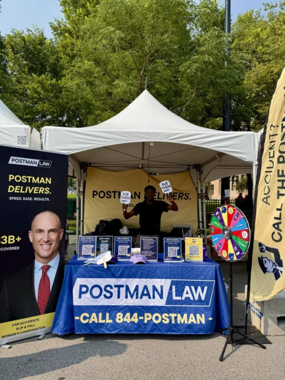 Un stand de Postman Law en un evento al aire libre muestra una pancarta, material promocional, una ruleta de premios y una persona que sostiene dos carteles de "Usted gana" bajo una carpa blanca.