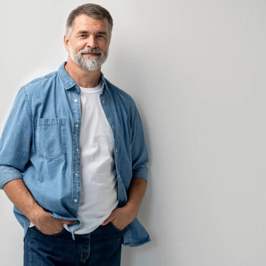 Middle-aged man with gray hair and beard, wearing a denim shirt over a white t-shirt and jeans, stands against a plain white background with hands in pockets.