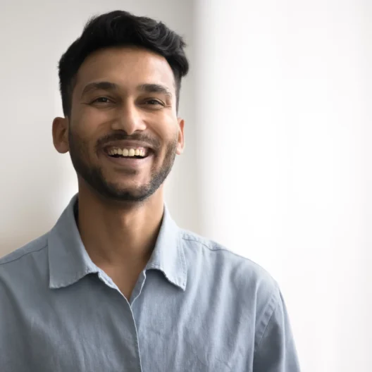 A man with short dark hair and a beard is smiling while standing indoors, wearing a light blue button-up shirt. The background is softly lit and out of focus.