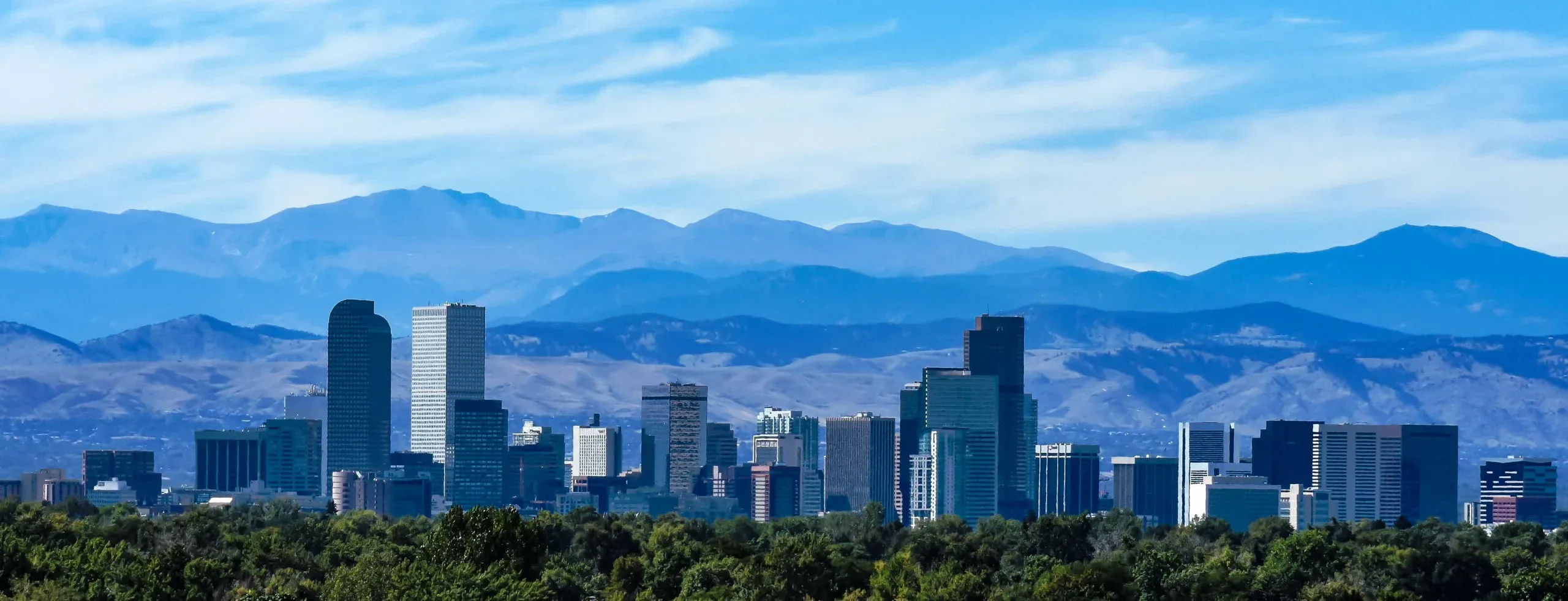 Horizonte de Denver con edificios altos en primer plano, árboles verdes debajo y montañas con un cielo azul al fondo.