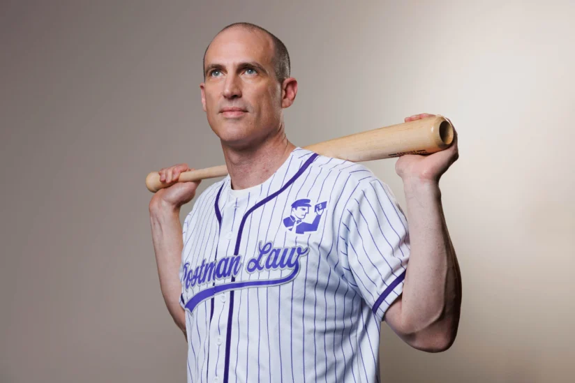 Un hombre con una camiseta de béisbol a rayas que sostiene un bate sobre los hombros está de pie sobre un fondo liso, mirando ligeramente hacia arriba.